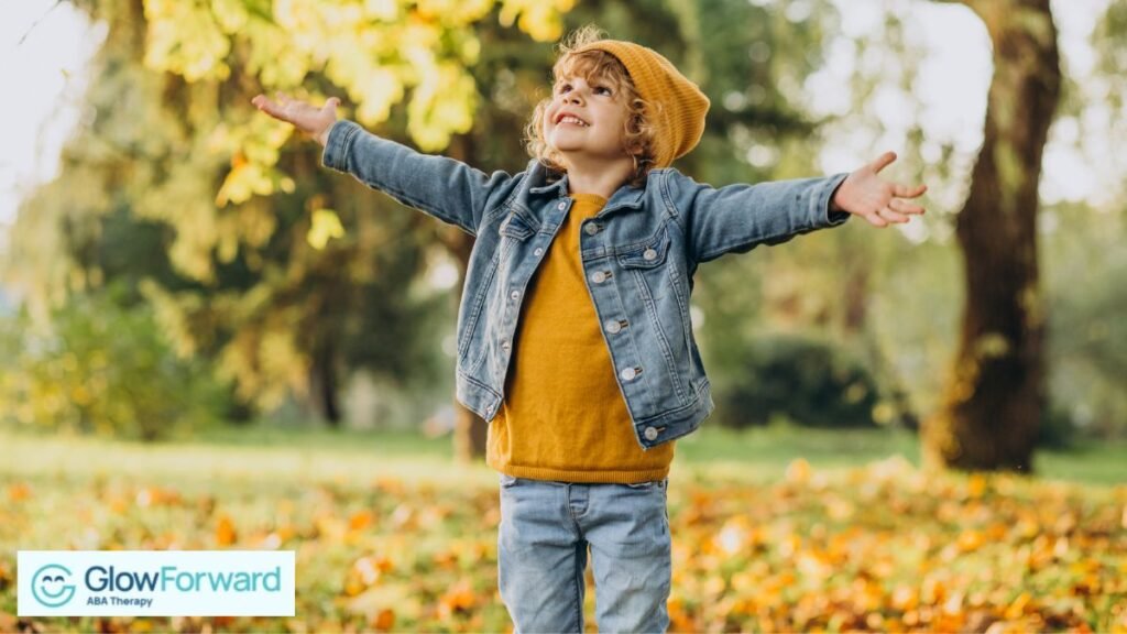 A blonde boy raises his arms toward the sky while standing in the middle of an autumn forest.