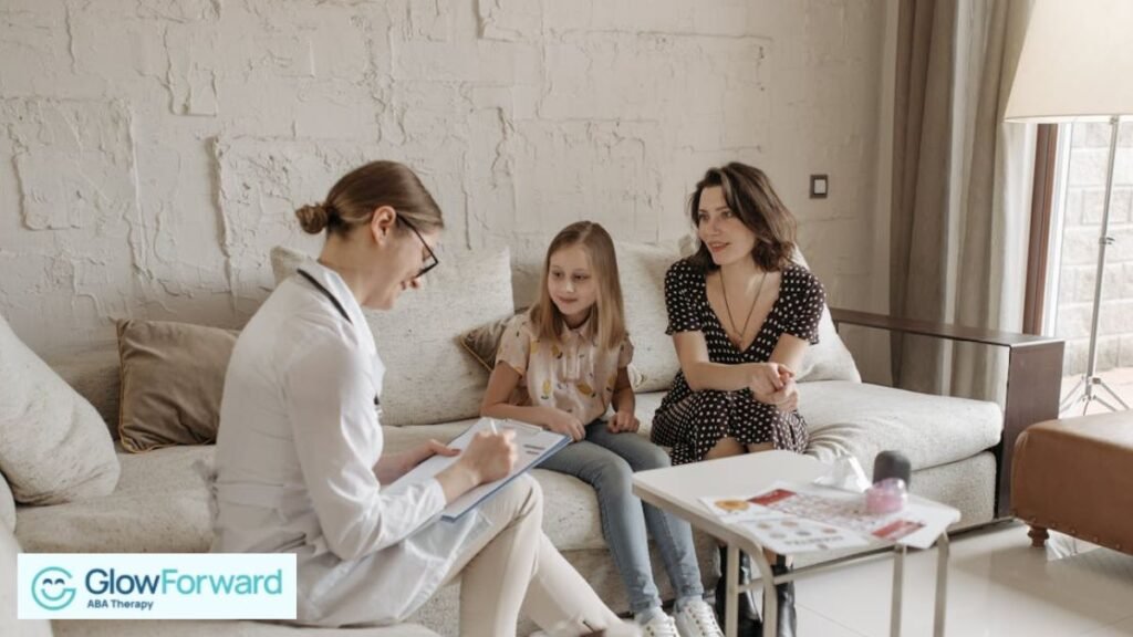 A female therapist looks down at her record board while listening to a mother discuss her daughter in a white therapy room.